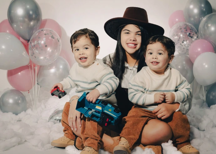 Sesión de Hermanos, una captura de tres hermanos compartiendo un momento de juego rodeados de globos y juguetes sobre una base que simula nubes. La imagen resalta la interacción natural en un entorno decorado de manera festiva.