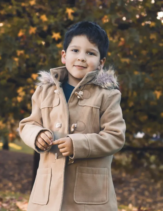 Retrato de medio cuerpo de un niño de pie, vistiendo un cálido abrigo color camel con botones de cuerno y capucha ribeteada de pelo, posando en un entorno de parque con árboles de fondo y hojas de color otoñal.