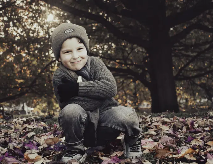 Retrato de cuerpo entero de un niño sonriente en cuclillas, vestido con un suéter de punto gris, guantes negros y vaqueros, rodeado de hojas secas moradas y marrones, con árboles oscuros al fondo y luz del sol filtrándose.