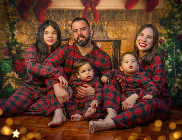 Fotografía de estudio de una familia de cinco miembros: padre, madre, una niña y dos niños. Visten pijamas navideños coordinados y están sentados juntos. La iluminación es suave y profesional, destacando sus expresiones felices en un entorno sin distracciones."
