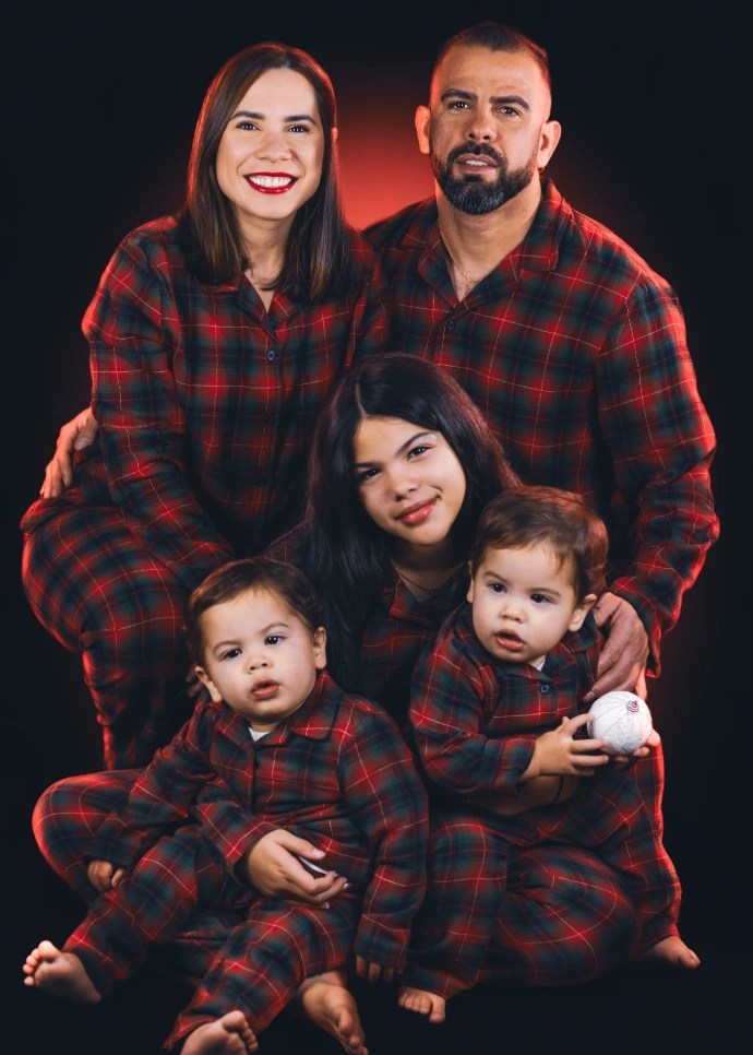 "Retrato de familia numerosa en estudio con fondo negro, y con una luz roja de fondo que se refleja en la ropa y cabello. padres y tres niños posan juntos, todos con pijamas navideños. Las expresiones son alegres y cálidas, capturando un momento familiar especial."