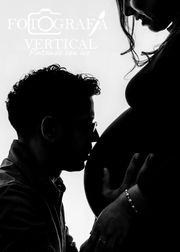 Silueta artística en blanco y negro de pareja capturando la conexión del embarazo a contraluz en estudio profesional, papá besando con delicadeza el vientre de la mamá