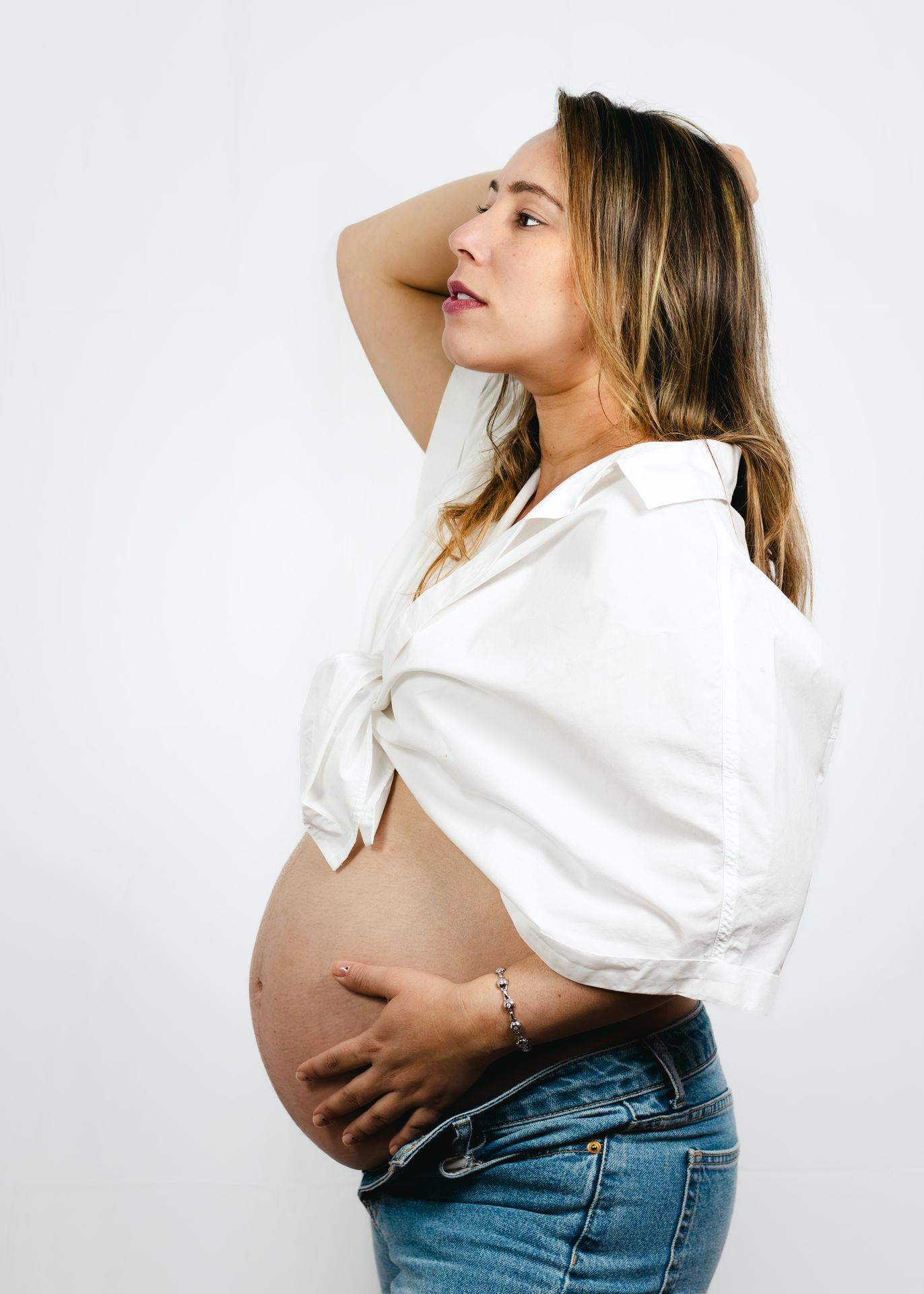 Sesión de fotos de maternidad estilo casual y natural con look moderno de camisa blanca y jeans en estudio.
