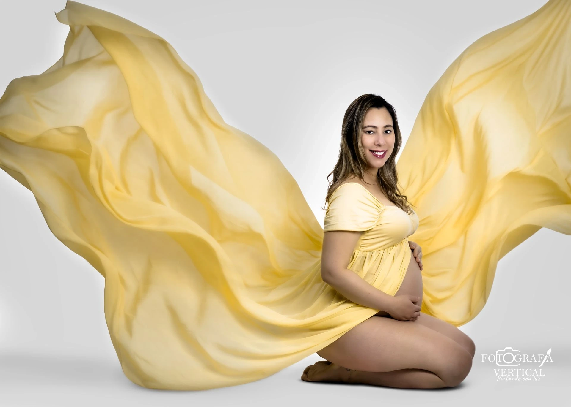 Fotografía artística de maternidad con vestido amarillo fluido y efecto de vestido volando con el viento para una sesión de fotos creativa.